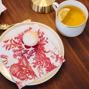 Une assiette blanche avec une pâtisserie en poudre et une serviette SERVIETTES 33X33 "LES 4 PARTIES DU MONDE" ROUGE GARANCE est posée à côté d'une tasse de thé au citron sur une table en bois, ainsi qu'une cuillère en or et un morceau de lampe. la marque Toile de Jouy