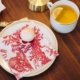 Une assiette blanche avec une pâtisserie en poudre et une serviette SERVIETTES 33X33 "LES 4 PARTIES DU MONDE" ROUGE GARANCE est posée à côté d'une tasse de thé au citron sur une table en bois, ainsi qu'une cuillère en or et un morceau de lampe. la marque Toile de Jouy