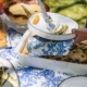 Une personne sert de la nourriture dans une assiette blanche avec SERVIETTES 33X33 "LES 4 PARTIES DU MONDE" BLEU DIRECTOIRE lors d'un repas en plein air avec divers plats sur une nappe bleue et blanche. la marque Toile de Jouy