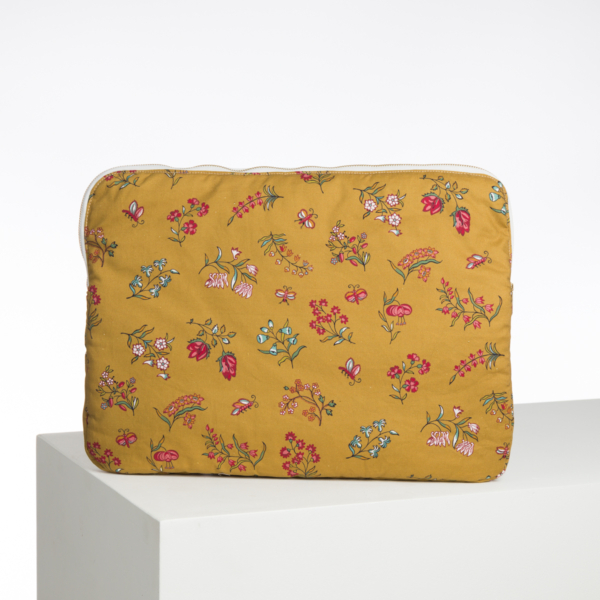 La XCOMM_CAPE DE PLUIE SEMIS DE FLEURS est une pochette en tissu jaune moutarde avec une fermeture éclair blanche et un motif floral coloré avec des fleurs roses, rouges, bleues et blanches, représenté sur une surface blanche sur un fond uni. la marque Toile de Jouy