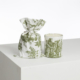 Le produit XCOMM_BOUGIE ET SON POCHON ROSIERE CUMIN présente une bougie blanche dans un support en verre avec des motifs de toile verte, placée à côté d'un sac à cordon en tissu assorti sur une surface blanche sur un fond uni. la marque Toile de Jouy