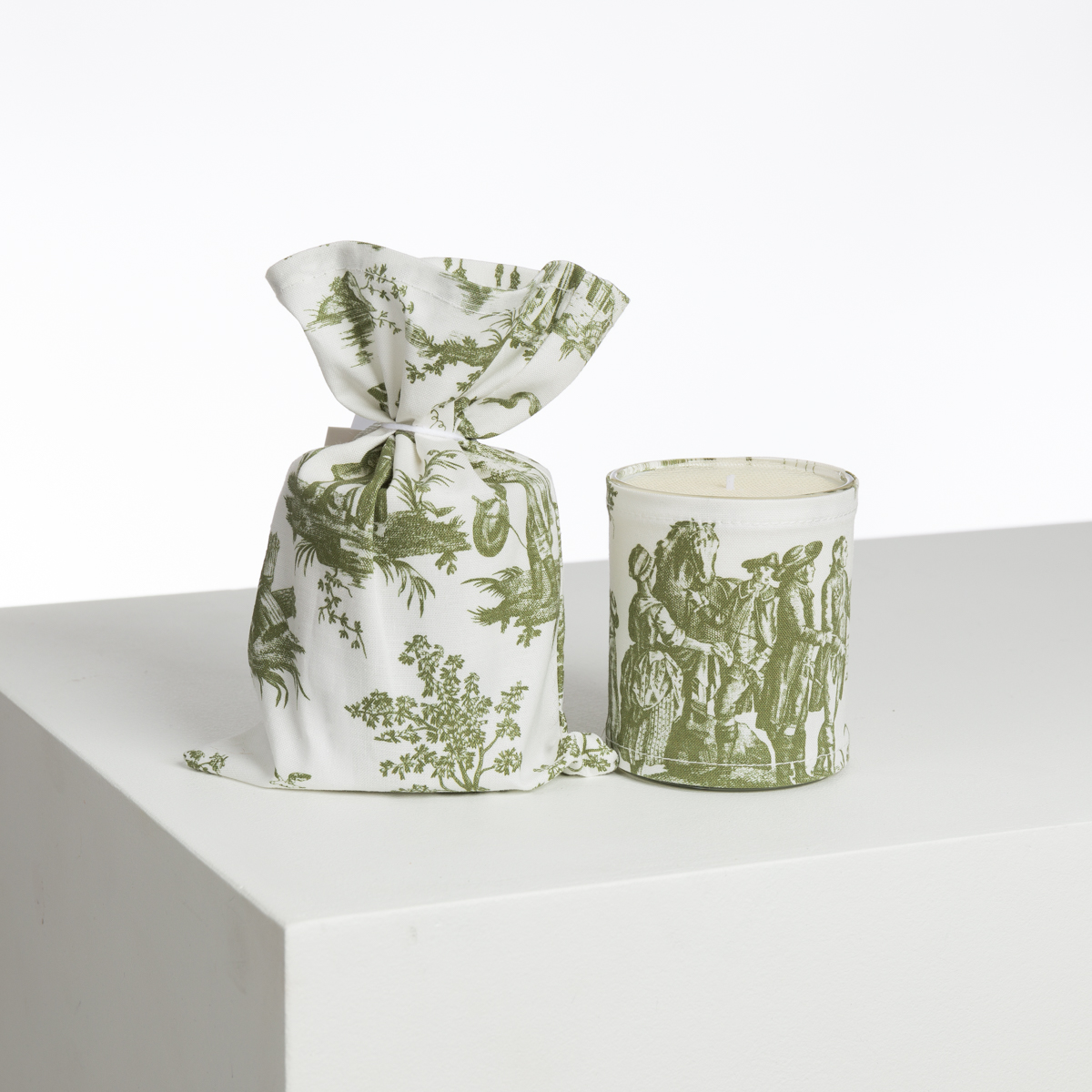 Le produit XCOMM_BOUGIE ET SON POCHON ROSIERE CUMIN présente une bougie blanche dans un support en verre avec des motifs de toile verte, placée à côté d'un sac à cordon en tissu assorti sur une surface blanche sur un fond uni. la marque Toile de Jouy