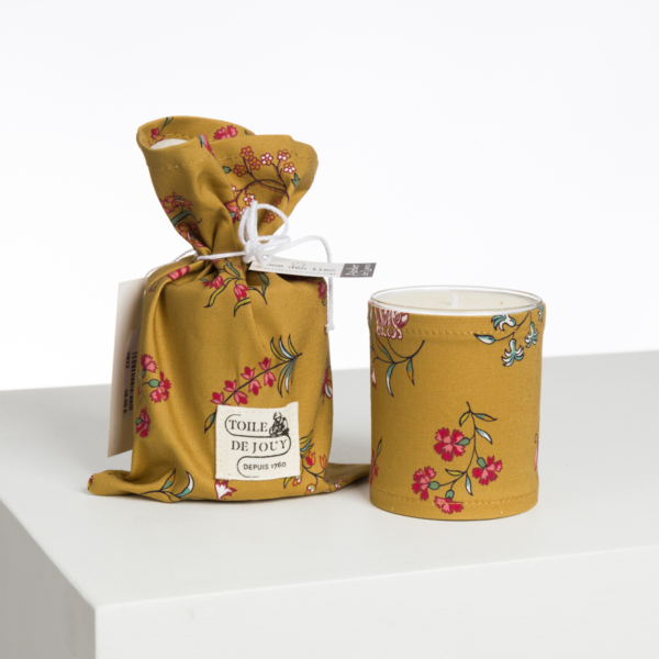 La XCOMM_CAPE DE PLUIE SEMIS DE FLEURS est une bougie jaune à motif floral, accompagnée d'une pochette en tissu et d'une étiquette blanche assorties, présentées sur un fond blanc uni. la marque Toile de Jouy