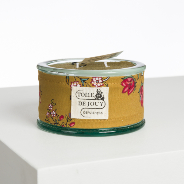 Une bougie ronde en verre enveloppée dans un tissu floral jaune, étiquetée "XCOMM_CAPE DE PLUIE SEMIS DE FLEURS", est posée sur une surface blanche sur un fond uni. la marque Toile de Jouy