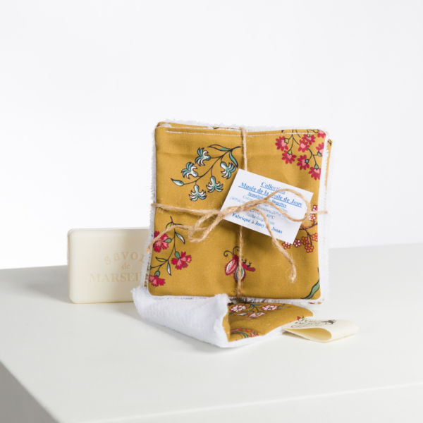 Une pile de tampons en coton réutilisables XCOMM_CAPE DE PLUIE SEMIS DE FLEURS jaunes à motifs floraux, attachés avec de la ficelle, se trouve à côté d'un savon blanc "Savon de Marseille" sur une surface blanche ; un tampon est déplié devant. la marque Toile de Jouy