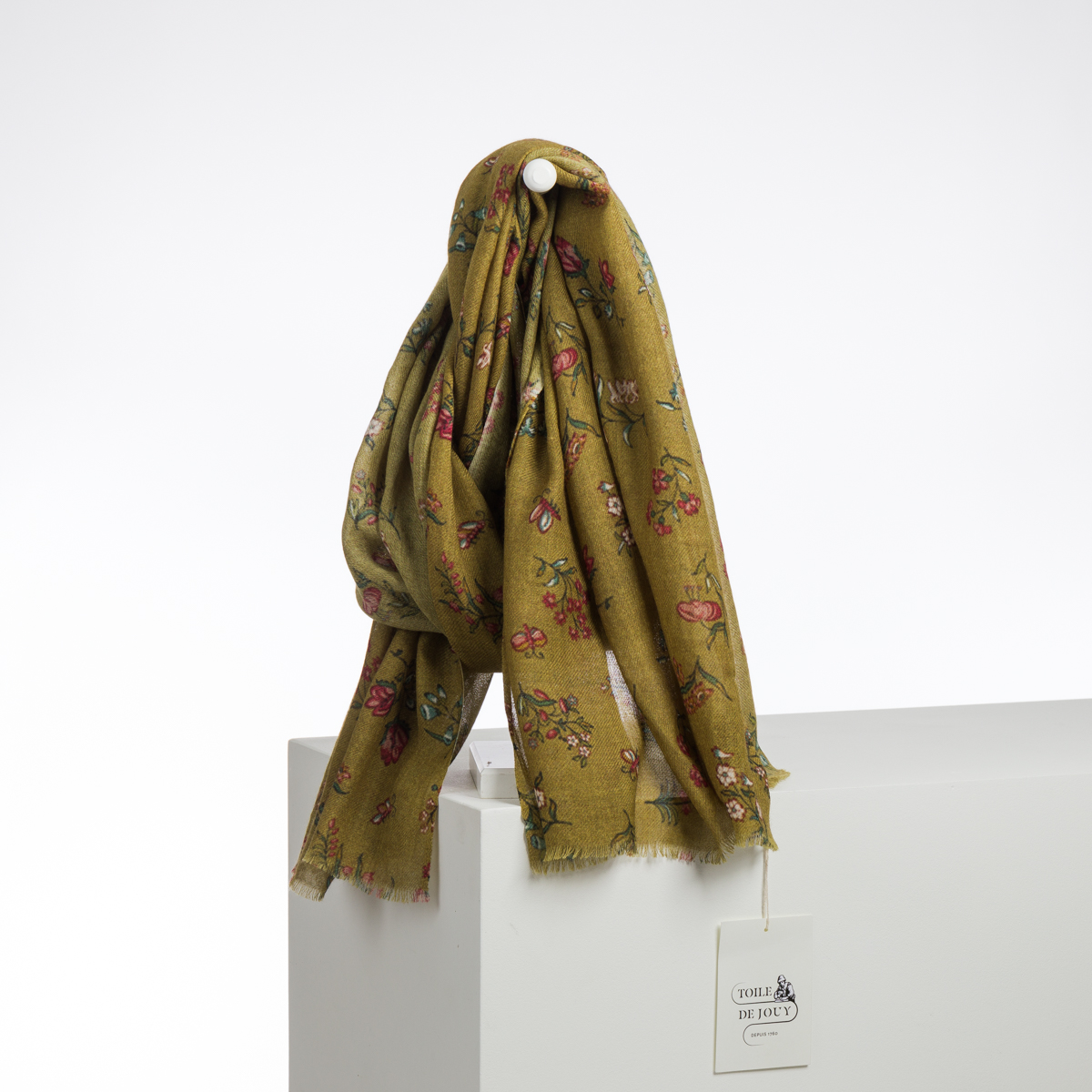 Une XCOMM_CAPE DE PLUIE SEMIS DE FLEURS jaune moutarde avec des motifs floraux roses, verts et rouges est suspendue à une cheville blanche au-dessus d'un bloc blanc. Une étiquette "Civell De Jolie" est attachée. Le fond est blanc uni. la marque Toile de Jouy