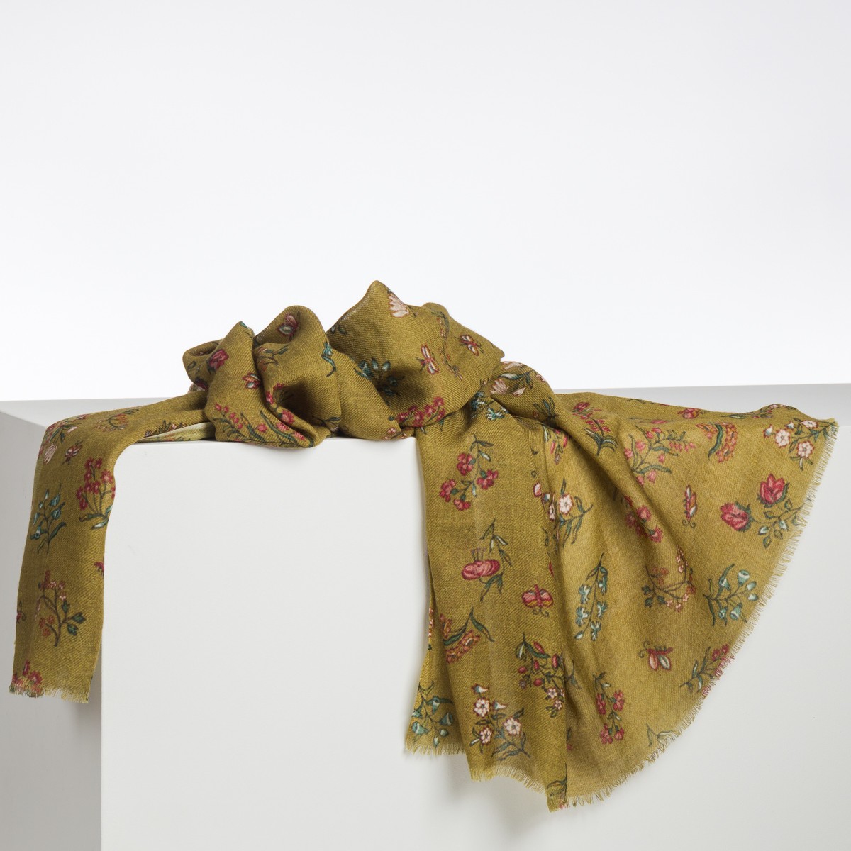 Le XCOMM_CAPE DE PLUIE SEMIS DE FLEURS est une douce écharpe vert olive avec un bord effiloché et un motif floral coloré, représentée drapée sur un bloc blanc sur un fond uni. la marque Toile de Jouy