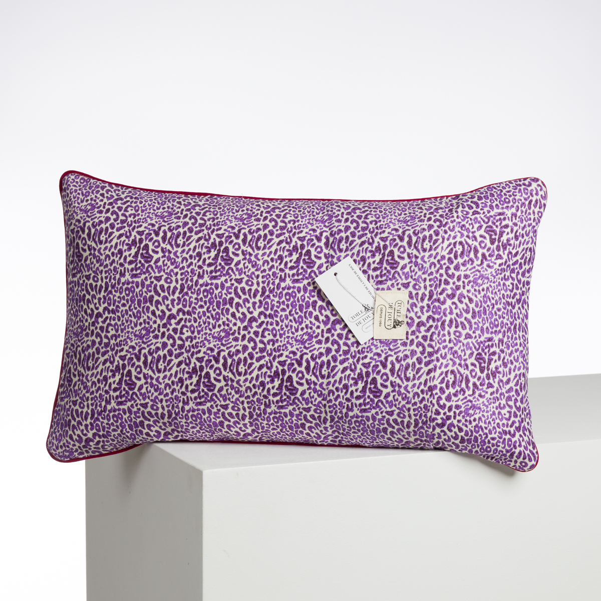 Le XCOMM_COUSSIN 30X50 ANANAS est un coussin rectangulaire avec un imprimé léopard violet et blanc, un passepoil rouge et une étiquette blanche, présenté sur une surface blanche sur un fond uni. la marque Toile de Jouy