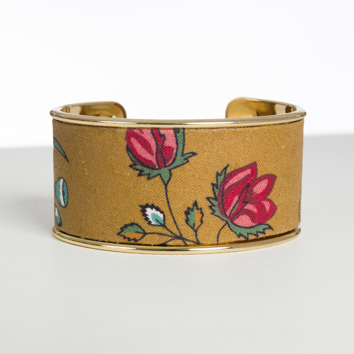 Le XCOMM_CAPE DE PLUIE SEMIS DE FLEURS est un bracelet manchette en or sur fond jaune moutarde, décoré de roses rouges et roses et de feuilles vertes, présenté sur une surface claire et neutre. la marque Toile de Jouy