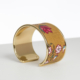 Le bracelet XCOMM_CAPE DE PLUIE SEMIS DE FLEURS est un bracelet manchette ouvert en or avec une incrustation jaune moutarde, présentant une fleur rouge et un petit motif floral blanc, sur une surface blanche et un fond uni. la marque Toile de Jouy