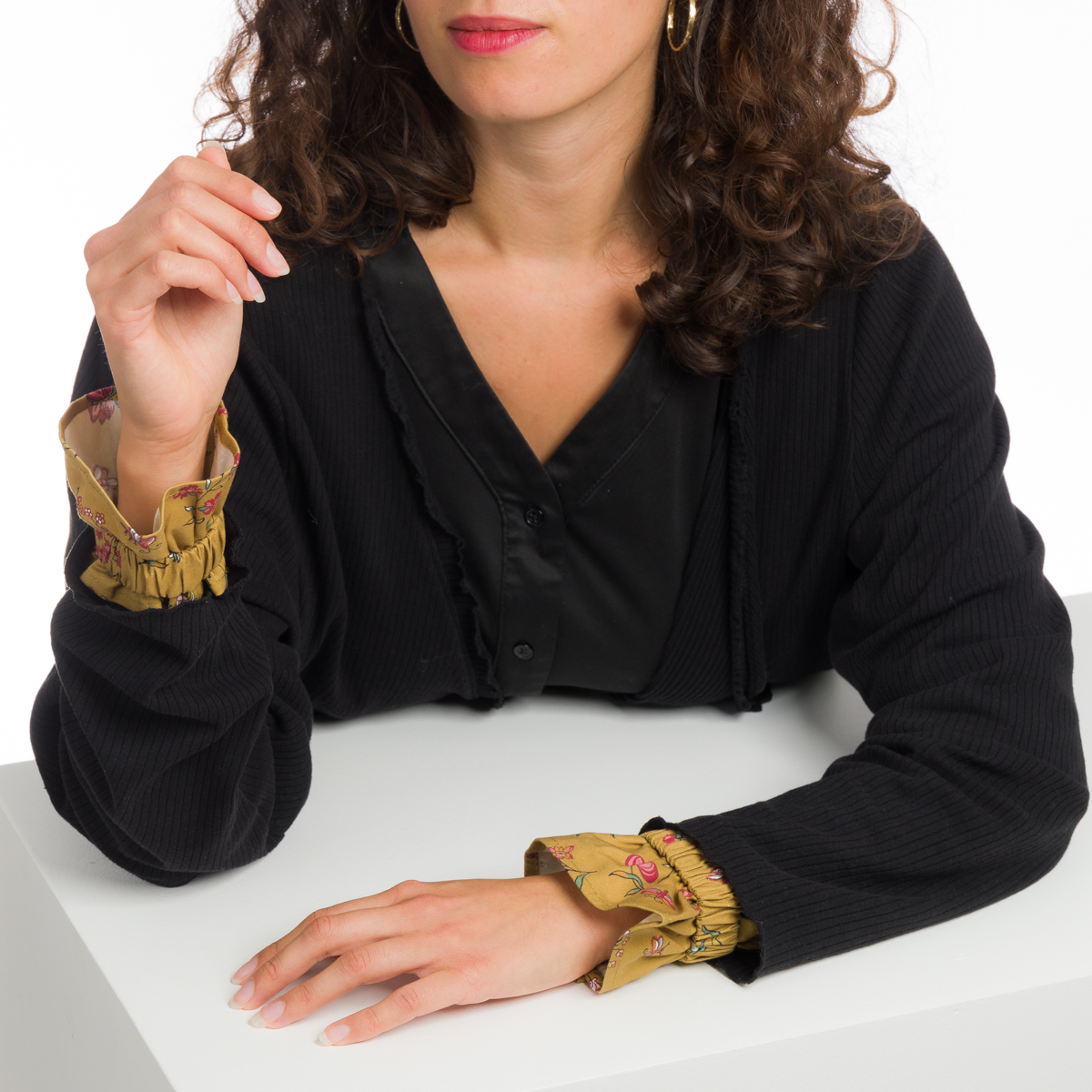 Une femme aux cheveux bouclés, avec des boucles d'oreilles dorées et un haut noir avec des poignets beiges à motifs, est assise à une table blanche et porte la XCOMM_CAPE DE PLUIE SEMIS DE FLEURS. Son visage est partiellement visible. la marque Toile de Jouy