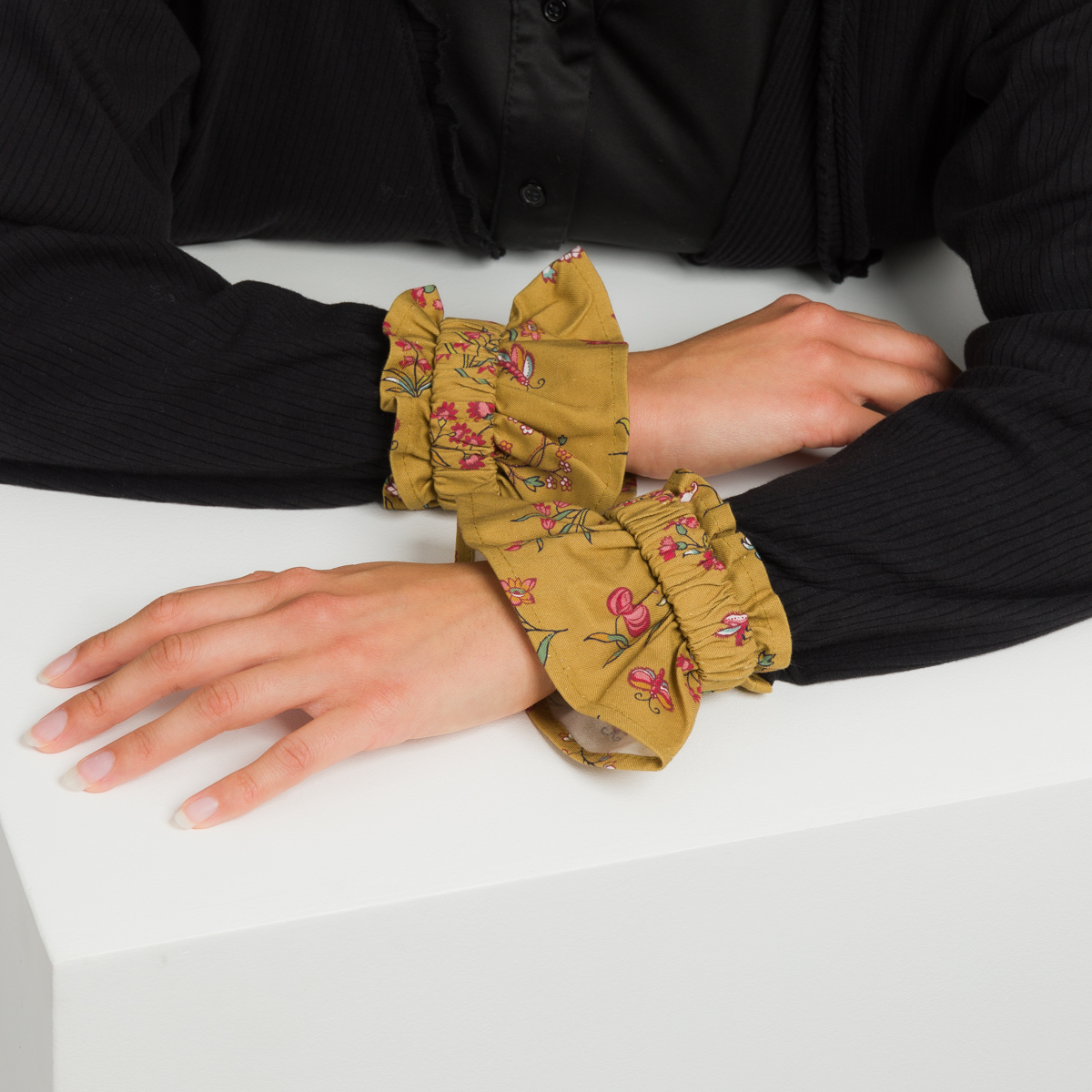 Une personne porte un haut noir à manches longues, les bras posés sur une surface blanche, les poignets ornés de la XCOMM_CAPE DE PLUIE SEMIS DE FLEURS - de larges manchettes jaunes à volants avec un motif floral rose. la marque Toile de Jouy