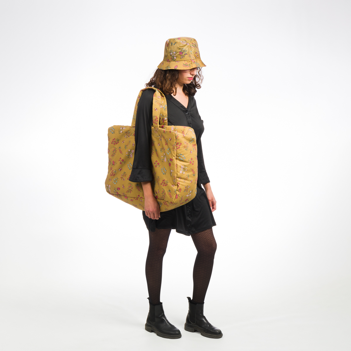 Vêtue d'une robe noire, de collants et de bottes, une personne se tient debout sur un fond blanc avec le chapeau à fleurs jaunes XCOMM_CAPE DE PLUIE SEMIS DE FLEURS et le sac surdimensionné assorti à l'épaule. la marque Toile de Jouy