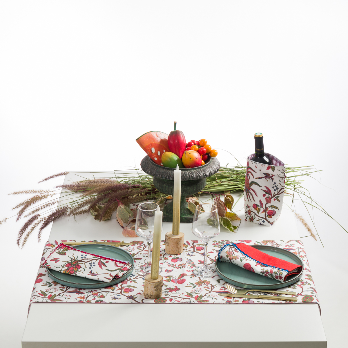 Une table à manger blanche comporte les sets de table XCOMM_SET DE TABLE/2 ANANAS, des serviettes, un porte-bouteille, deux assiettes, des bougies, des verres à vin et un centre de table composé de fruits et de feuilles d'automne. la marque Toile de Jouy