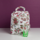 Le XCOMM_VANITY ANANAS, un sac en tissu blanc à motifs floraux avec poignée et fermeture éclair, repose sur une surface blanche devant un mur violet. À côté, une petite bouteille en verre vert avec un bouchon transparent. la marque Toile de Jouy