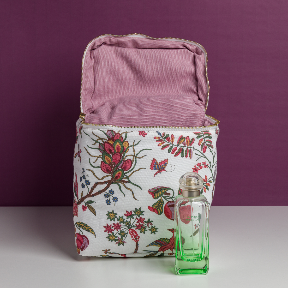 Le sac XCOMM_VANITY ANANAS, un sac en tissu à motifs floraux avec une doublure rose, est présenté ouvert sur une surface blanche. Devant lui se trouve un flacon de parfum en verre transparent rempli d'un liquide vert sur un fond violet uni. la marque Toile de Jouy