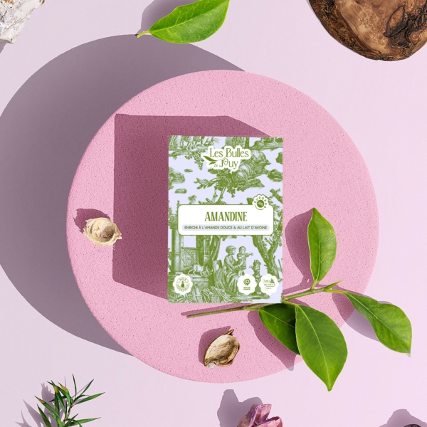Une boîte de savon Les Bulles de Juby "Amandine" est posée sur une plate-forme ronde et rose, entourée de feuilles vertes, de fleurs séchées, d'une planche en bois et de rochers, le tout sur un fond rose pâle. la marque Toile de Jouy