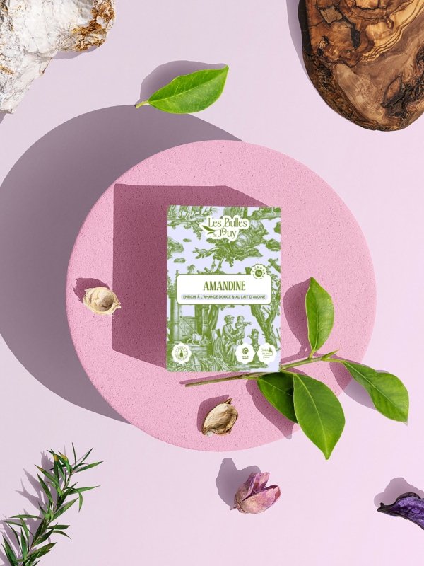 Une boîte de savon Les Bulles de Juby "Amandine" est posée sur une plate-forme ronde et rose, entourée de feuilles vertes, de fleurs séchées, d'une planche en bois et de rochers, le tout sur un fond rose pâle. la marque Toile de Jouy