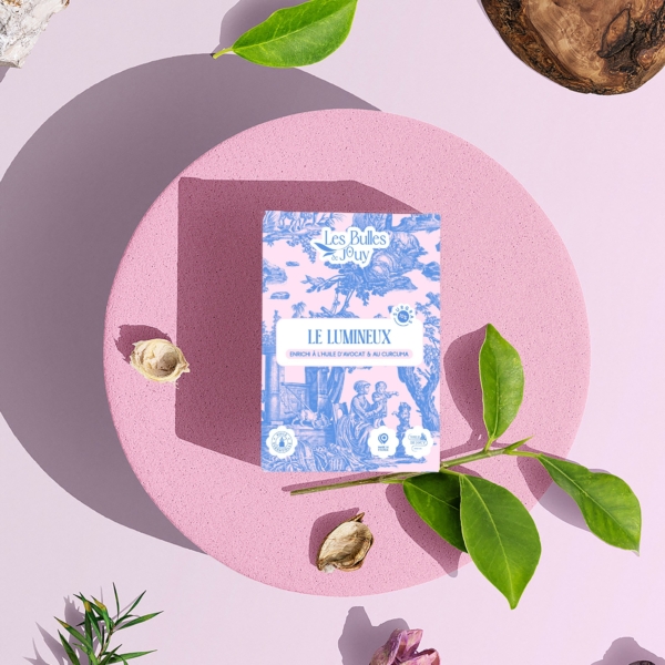 Une boîte de "Bulles de Joÿ Le Lumineux" est posée sur une plate-forme circulaire rose, entourée de feuilles vertes, de petites fleurs et de textures en bois sur un fond violet clair. la marque Toile de Jouy