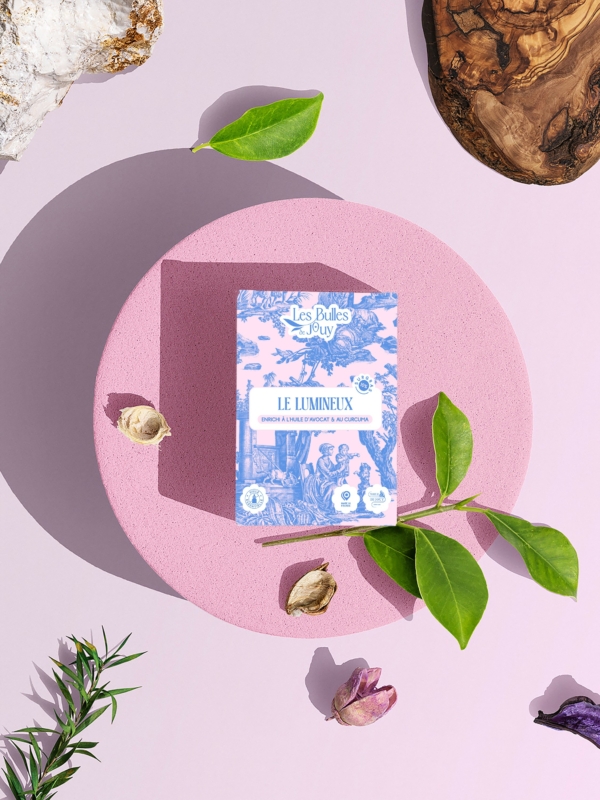 Une boîte de "Bulles de Joÿ Le Lumineux" est posée sur une plate-forme circulaire rose, entourée de feuilles vertes, de petites fleurs et de textures en bois sur un fond violet clair. la marque Toile de Jouy