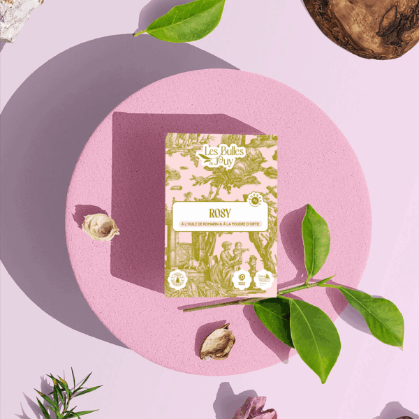Une boîte de savon rosé "Les Bulles de Joy" est posée sur une plate-forme ronde et rose, entourée de feuilles vertes, de fleurs séchées, d'une planche en bois et de pierres, sur un fond rose pâle. la marque Toile de Jouy