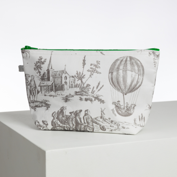Le XCOMM_LUNCH BAG ESCARPOLETTE ROUGE est une pochette blanche à fermeture éclair avec une doublure verte, présentant des illustrations noir et blanc de style vintage de personnes, d'une église, d'arbres et d'une montgolfière, sur une surface blanche et un fond uni. la marque Toile de Jouy