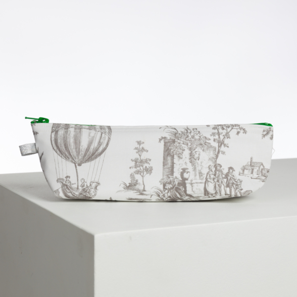 Le XCOMM_LUNCH BAG ESCARPOLETTE ROUGE est une pochette blanche à fermeture éclair verte, décorée d'illustrations grises de style vintage représentant des personnes, des arbres, des bâtiments et une montgolfière. Il repose sur une surface blanche sur un fond uni. la marque Toile de Jouy