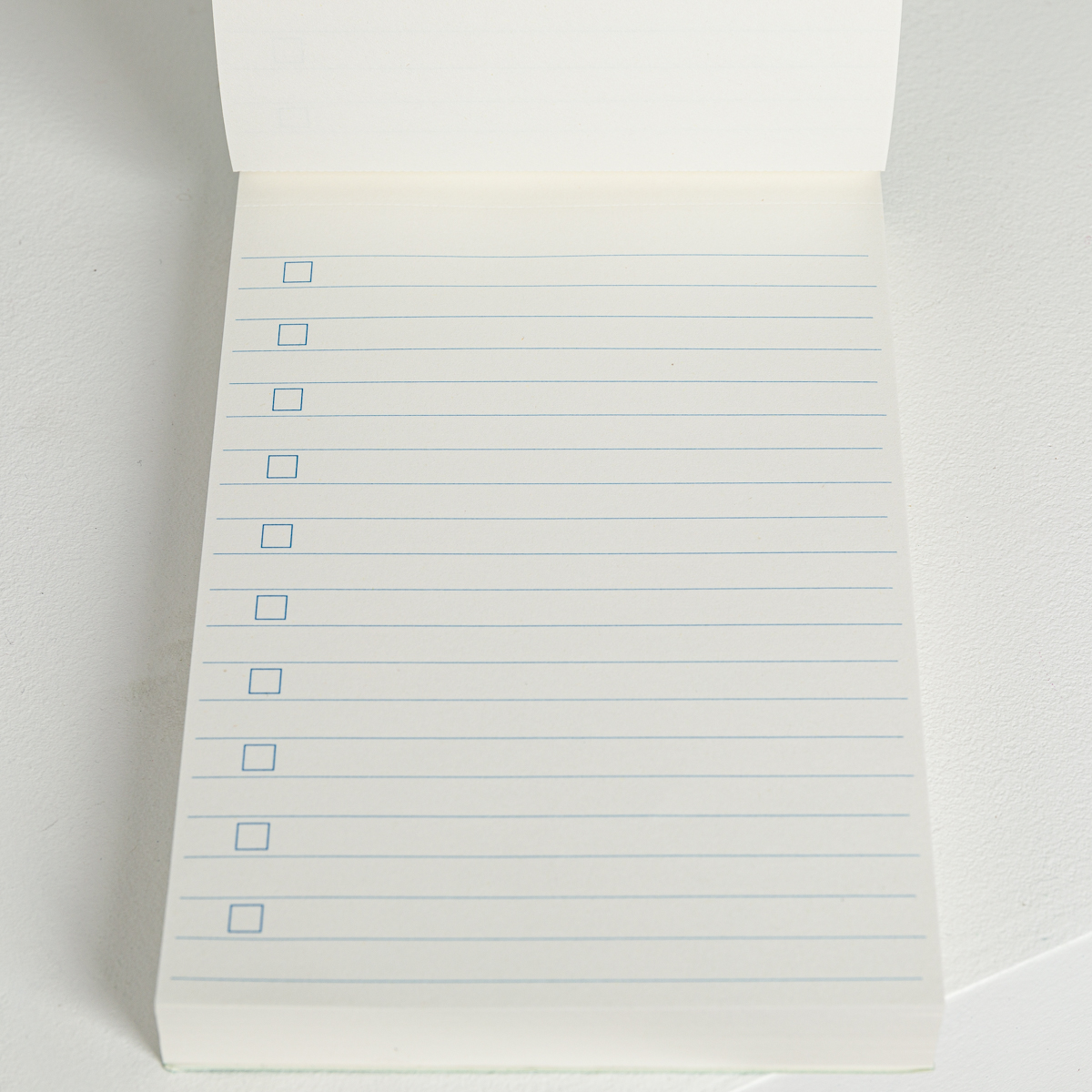 La liste XCOMM_EVERYDAY COURONNEMENT VERT PRAIRIE est un bloc-notes ouvert avec des lignes horizontales bleues et des cases à cocher vides sur le côté gauche de chaque ligne, affichées sur une surface blanche. la marque Toile de Jouy