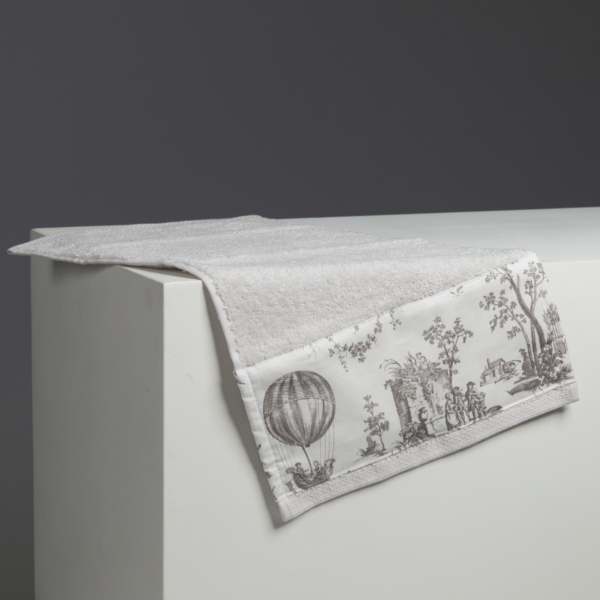 Le XCOMM_LUNCH BAG ESCARPOLETTE ROUGE est présenté drapé sur une surface rectangulaire blanche, avec des illustrations de style vintage telles qu'une montgolfière et des personnes le long de son bord, sur un fond gris foncé. la marque Toile de Jouy