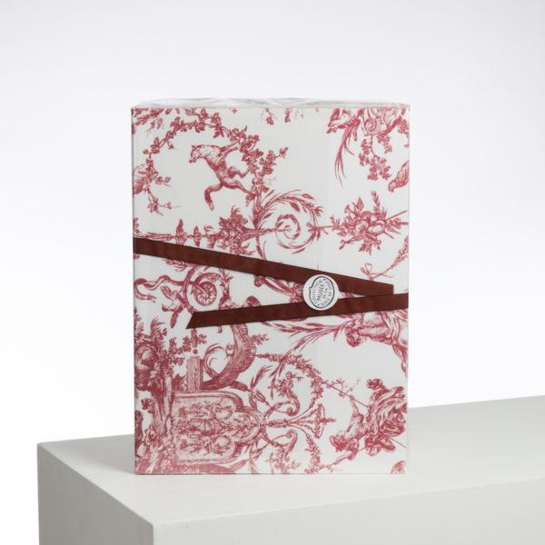 Le XCOMM_LUNCH BAG ESCARPOLETTE ROUGE est un sac rectangulaire enveloppé dans du papier blanc avec des motifs floraux et pastoraux rouges, noué avec un ruban marron et une étiquette ronde, présenté sur une surface blanche sur un fond uni. la marque Toile de Jouy