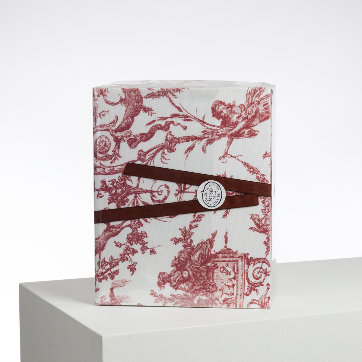 Le XCOMM_LUNCH BAG ESCARPOLETTE ROUGE, emballé dans un papier blanc avec des illustrations rouges de style vintage et noué avec un ruban rouge, est posé sur une surface blanche sur un fond uni. la marque Toile de Jouy