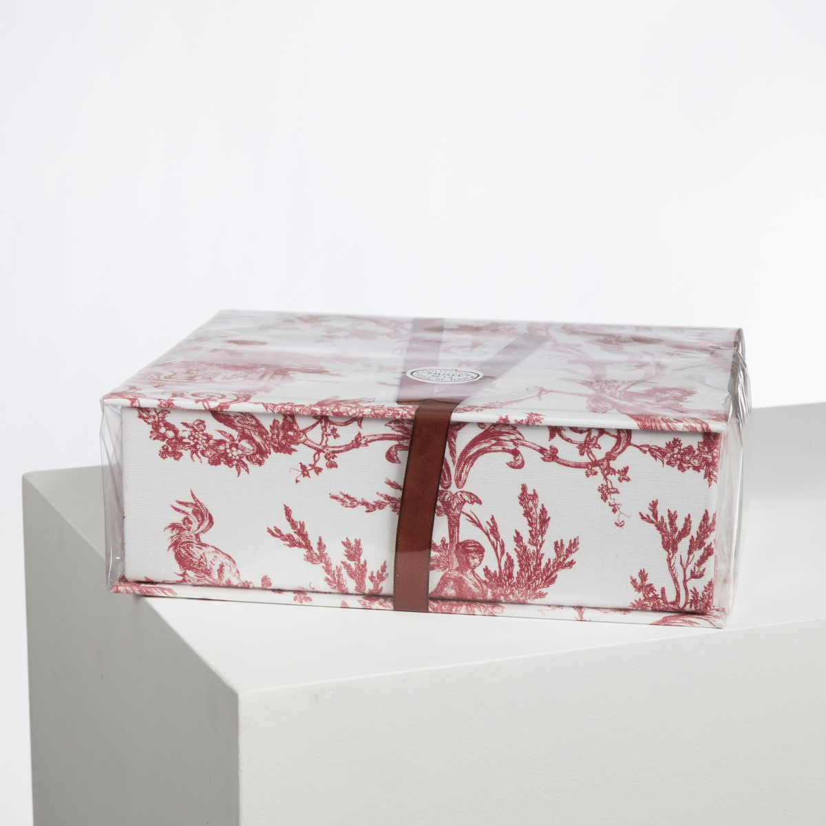 Un XCOMM_LUNCH BAG ESCARPOLETTE ROUGE, orné de motifs floraux et animaliers rouges sur fond blanc et noué par un ruban bordeaux, est posé sur une surface blanche sur un fond blanc uni. la marque Toile de Jouy