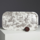 Le XCOMM_LUNCH BAG ESCARPOLETTE ROUGE, qui présente des illustrations pastorales en noir et blanc, est posé sur un piédestal blanc devant lequel se trouve un objet sphérique foncé et texturé, sur fond gris. la marque Toile de Jouy
