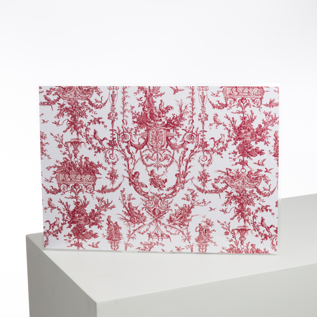 Le XCOMM_LUNCH BAG ESCARPOLETTE ROUGE est présenté à la verticale sur un piédestal géométrique blanc, mettant en valeur son motif de toile rouge et blanche orné de motifs et de figures classiques complexes sur un fond uni. la marque Toile de Jouy