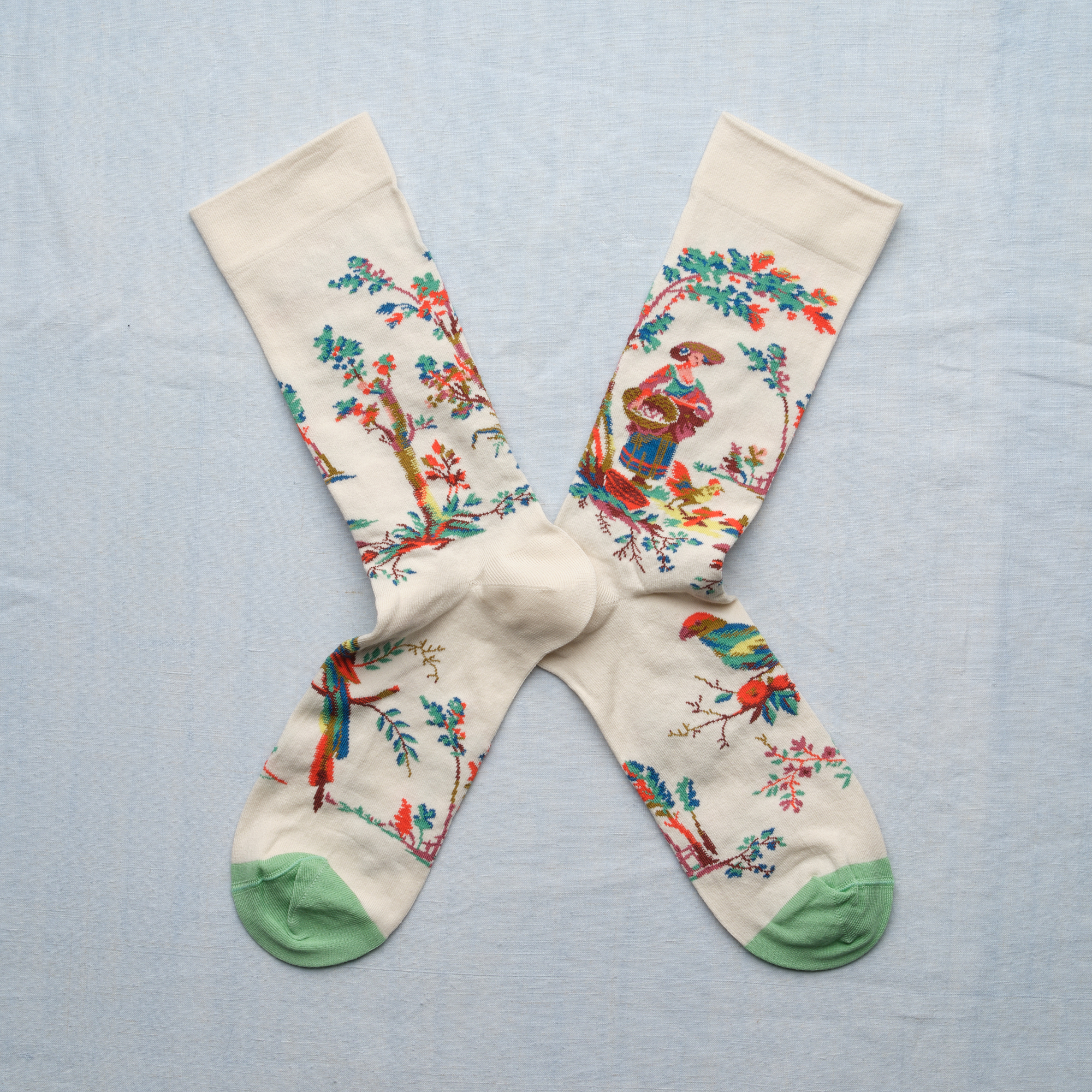 Les XCOMM_CHAUSETTES PLAISIRS DE LA CAMPAGNE 36/38 sont des chaussettes blanches avec des orteils vert menthe, des motifs floraux colorés et un personnage traditionnel, représenté croisé sur un fond bleu clair. la marque Toile de Jouy