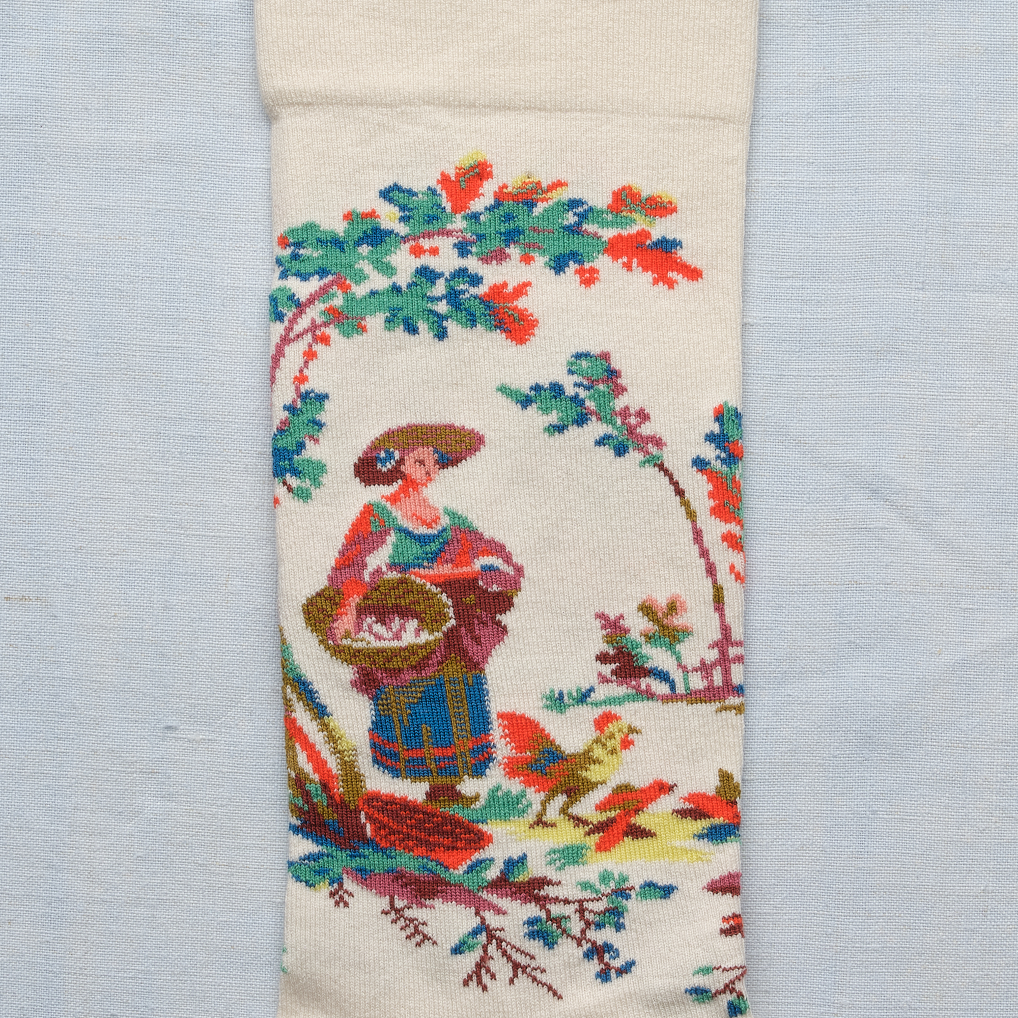 Les XCOMM_CHAUSETTES PLAISIRS DE LA CAMPAGNE 36/38 présentent un tissu de couleur crème avec des broderies colorées représentant une femme et un enfant sous une arche feuillue, des motifs floraux et végétaux, le tout sur un fond bleu clair. la marque Toile de Jouy