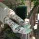 Se détendant en plein air au soleil, une personne portant un pantalon blanc soutient ses pieds - ornés de XCOMM_CHAUSETTES PLAISIRS DE LA CAMPAGNE 36/38, avec des oiseaux colorés et des motifs floraux - sur une clôture rustique en bois au milieu d'un environnement verdoyant. la marque Toile de Jouy