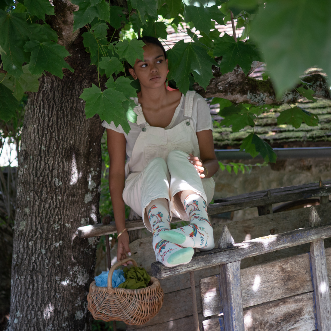 Une jeune femme en salopette blanche est assise sur une échelle en bois sous un arbre, portant XCOMM_CHAUSETTES PLAISIRS DE LA CAMPAGNE 36/38 des chaussettes colorées ; ses jambes sont croisées, et un panier avec du tissu repose à proximité sur les barreaux. la marque Toile de Jouy