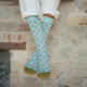 Quelqu'un porte XCOMM_CHAUSETTES PLAISIRS DE LA CAMPAGNE 36/38 - des chaussettes bleues claires à motifs - avec un pantalon blanc retroussé, assis les jambes croisées contre un mur de pierre rustique. la marque Toile de Jouy