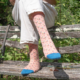 Une personne est assise en plein air sur un banc usé par les intempéries, entouré de verdure, et porte XCOMM_CHAUSETTES PLAISIRS DE LA CAMPAGNE 36/38 : des chaussettes rose pâle avec des orteils bleus et un petit motif floral. On ne voit que le bas des jambes dans un pantalon blanc. la marque Toile de Jouy