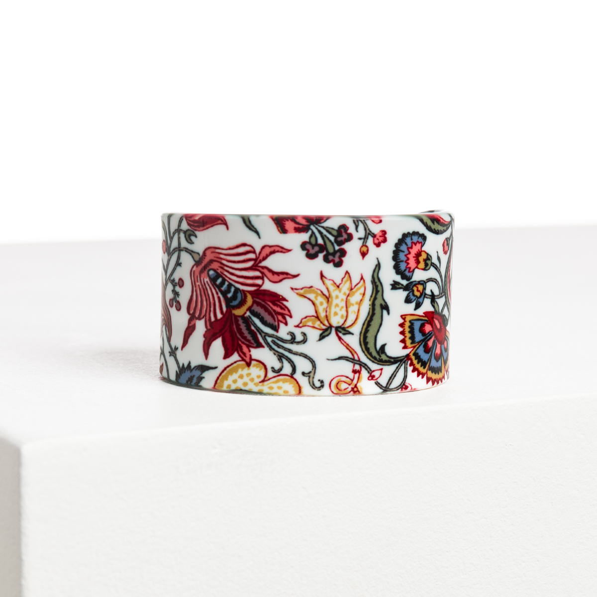 L'ABAT-JOUR SUR COMMANDE est un rond de serviette en céramique blanche présentant des motifs floraux complexes rouges, jaunes et bleus sur une surface blanche et un fond uni. la marque Toile de Jouy