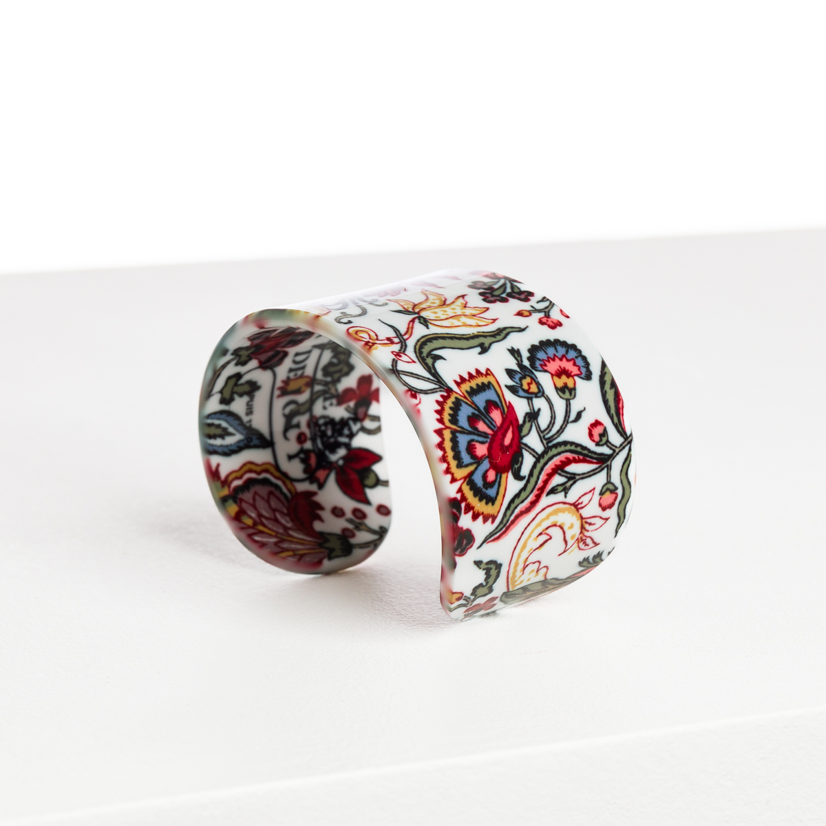 ABAT-JOUR SUR COMMANDE : Un bracelet large et incurvé avec une finition brillante et un motif floral et de feuilles vibrant en rouge, vert, jaune et bleu sur blanc, présenté sur une surface blanche. la marque Toile de Jouy