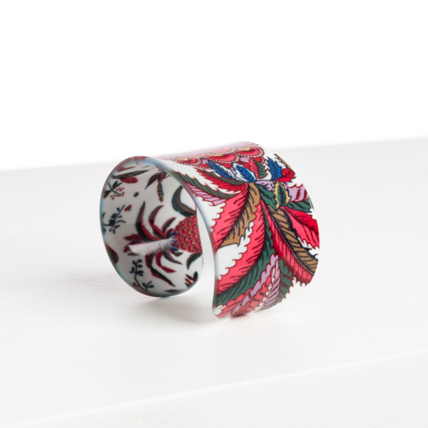 Le bracelet ABAT-JOUR SUR COMMANDE est un bracelet manchette large et brillant qui présente un motif floral et feuillu vibrant en rouge, rose, bleu et vert sur fond blanc, présenté debout sur une surface blanche. la marque Toile de Jouy