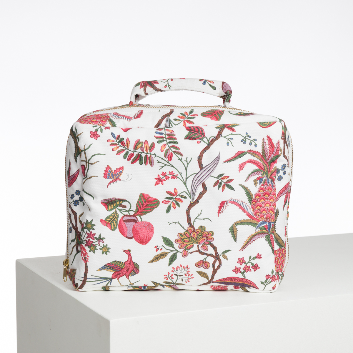 L'ABAT-JOUR SUR COMMANDE est une trousse à cosmétiques carrée de couleur blanche, ornée d'un motif floral et d'oiseaux colorés en rose, rouge et vert. Il est doté d'une fermeture à glissière et d'une petite poignée supérieure. Il est présenté sur une surface blanche sur un fond uni. la marque Toile de Jouy