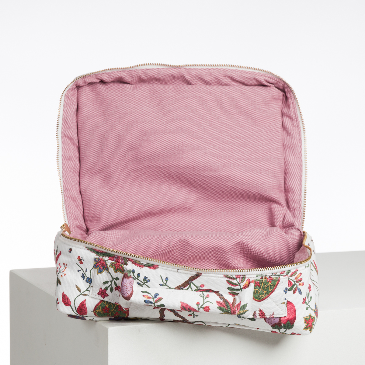 L'ABAT-JOUR SUR COMMANDE est une pochette en tissu avec un intérieur rose et une ouverture zippée, avec un extérieur blanc orné de motifs floraux et d'oiseaux colorés. Elle est présentée ouverte et vide sur une surface blanche. la marque Toile de Jouy