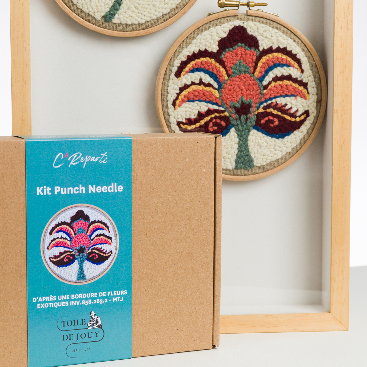 La boîte du kit ABAT-JOUR SUR COMMANDE, ornée d'une étiquette florale brodée, est placée devant un cadre en bois qui présente l'œuvre d'art à l'aiguille poinçonnée, avec un motif floral symétrique et vibrant. la marque Toile de Jouy