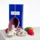 Une paire de pantoufles florales ABAT-JOUR SUR COMMANDE est présentée devant une boîte bleue de L'Atelier Charentaises, avec à côté des décorations en bois de fruits et légumes sur une surface blanche. la marque Toile de Jouy