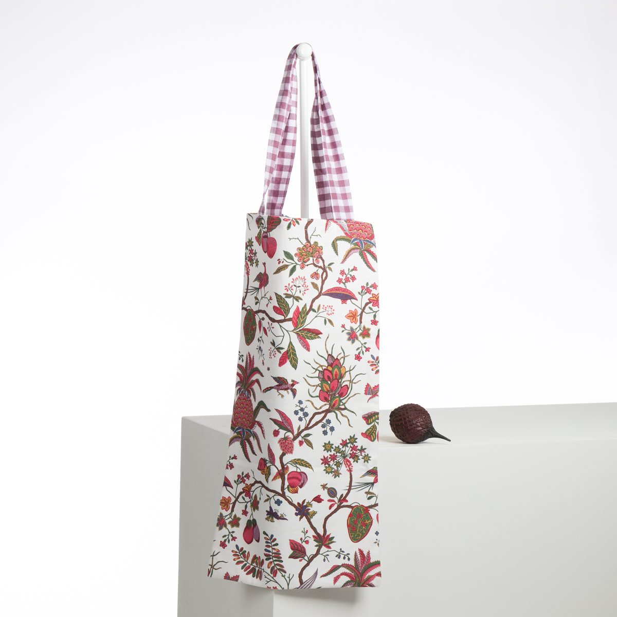 L'ABAT-JOUR SUR COMMANDE, un sac fourre-tout fleuri avec des motifs roses et verts et une poignée en vichy rose et blanc, est suspendu sur un rebord blanc à côté d'une petite betterave violet foncé sur fond blanc. la marque Toile de Jouy