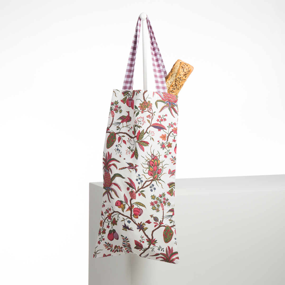 L'ABAT-JOUR SUR COMMANDE, un sac fourre-tout réutilisable en tissu avec un imprimé floral vif et des poignées à carreaux roses, est suspendu à un crochet blanc. Une baguette de pain dépasse du haut du sac sur un fond blanc uni. la marque Toile de Jouy