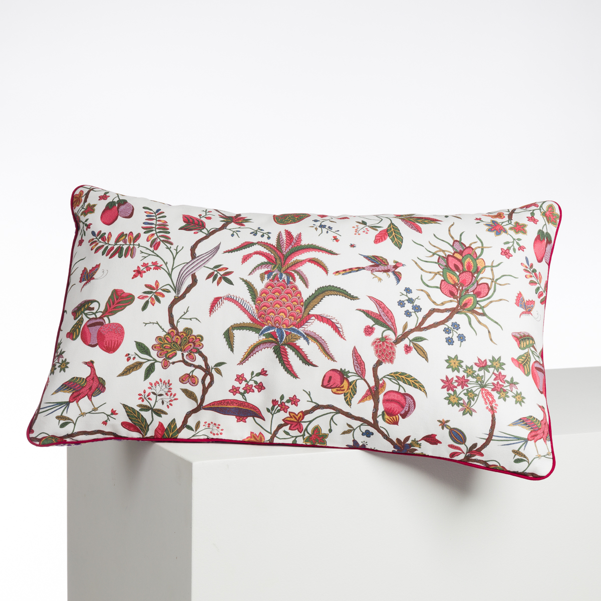 ABAT-JOUR SUR COMMANDE est un coussin rectangulaire à fond blanc, décoré de motifs floraux et fruitiers roses, verts et rouges, avec un liseré rose foncé sur les bords. Il est présenté reposant sur une surface blanche. la marque Toile de Jouy